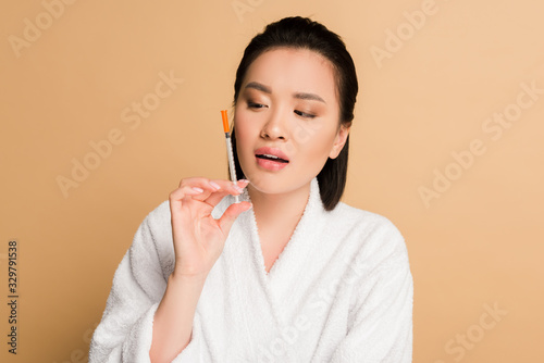 beautiful asian woman in bathrobe looking at syringe for beauty injection on beige background