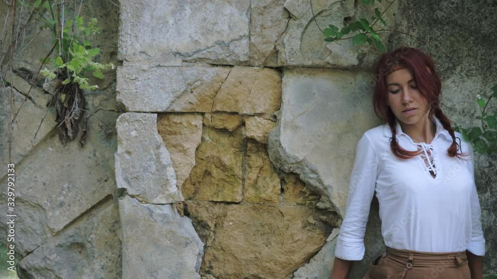 A beautiful red-haired girl in medieval rustic clothes walks over a cliff along a ledge along the wall of the fortress. A beautiful red-haired girl in medieval rustic clothes walks over a cliff along a ledge along the wall of the fortress.