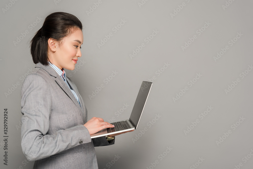 side view of smiling young businesswoman in suit using laptop on grey background side view of smiling young businesswoman in suit using laptop on grey background