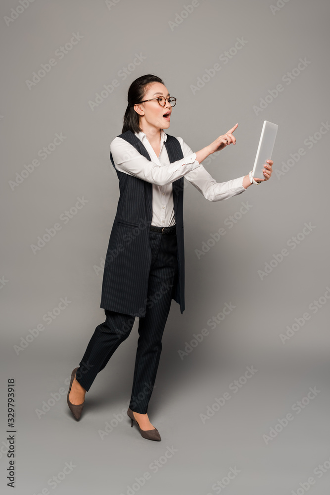 young businesswoman in eyeglasses with open mouth pointing with finger at digital tablet on grey background