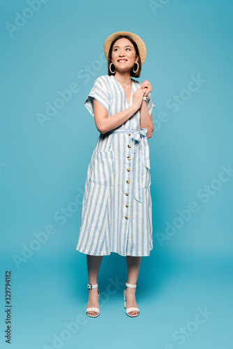 smiling brunette asian girl in striped dress and straw hat on blue background