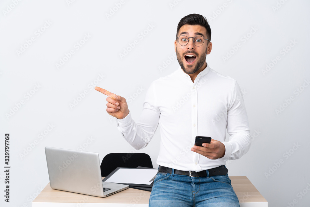 Young businessman holding a mobile phone surprised and pointing finger to the side Young businessman holding a mobile phone surprised and pointing finger to the side
