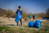 young blond hair woman with her sons in blue gloves cleaning the river bank from plastic and garbage in big blue trash bags on a sunny day in early spring