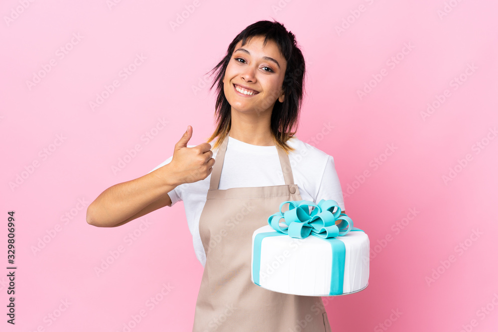 Uruguayan Pastry chef holding a big cake over isolated pink background giving a thumbs up gesture Uruguayan Pastry chef holding a big cake over isolated pink background giving a thumbs up gesture