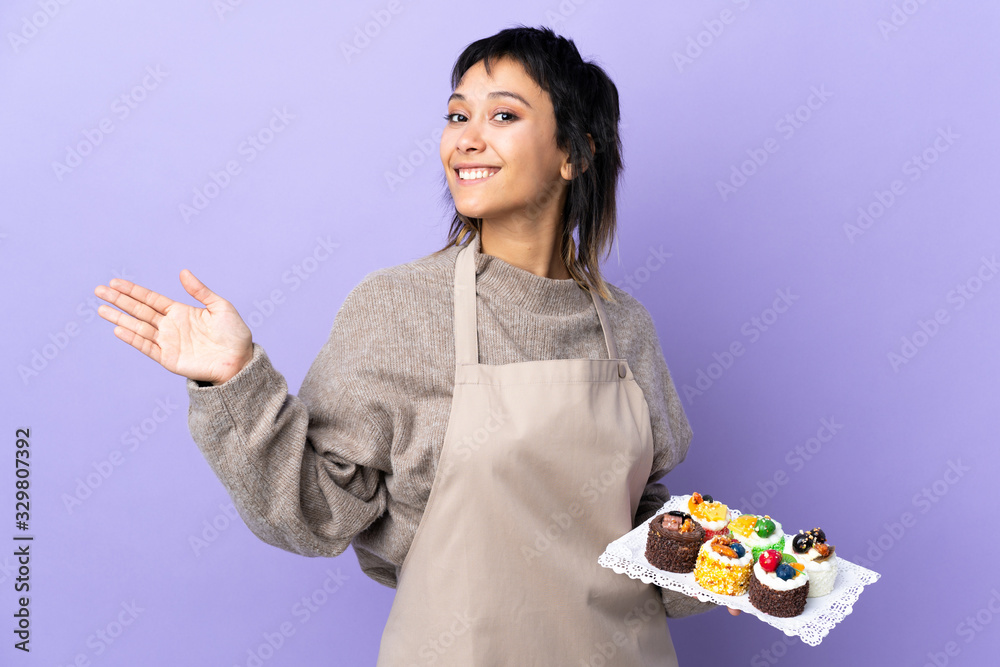 Young Uruguayan woman holding lots of different mini cakes over isolated purple background extending hands to the side for inviting to come Young Uruguayan woman holding lots of different mini cakes over isolated purple background extending hands to the side for inviting to come