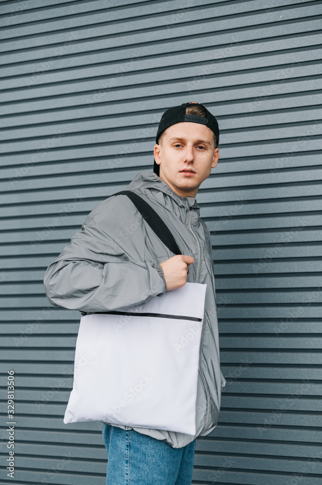 Portrait of attractive young man in stylish streetwear with white eco bag on shoulder posing on gray background with serious face. Guy with stylish reusable eco bag. Vertical. Portrait of attractive young man in stylish streetwear with white eco bag on shoulder posing on gray background with serious face. Guy with stylish reusable eco bag. Vertical.