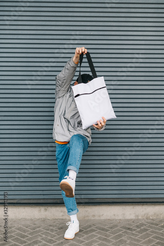Funny young man in trendy streetwear having fun with eco bag in his hands on gray wall background, vertical photo. An expressive guy holds a reusable shopping bag in his hand and lifts his leg.