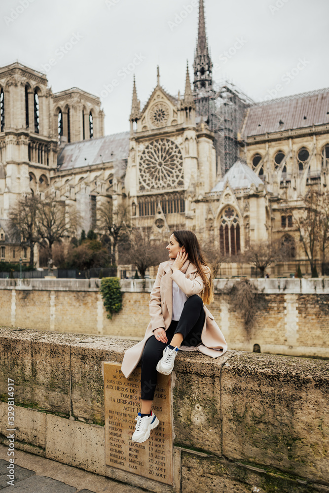Happy stylish girl having fun near Notre-Dame cathedral in Paris, France. Tourist enjoying their vacation in France. Young smiling woman tourist sitting in front of the famous Notre Dame cathedral.