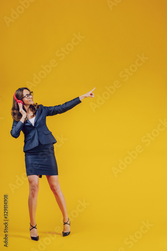 European woman in high spirit points away with thumb, feels entertained and satisfied, listens radio broadcasting with modern headphones connected to telephone, checks sound, shows free space