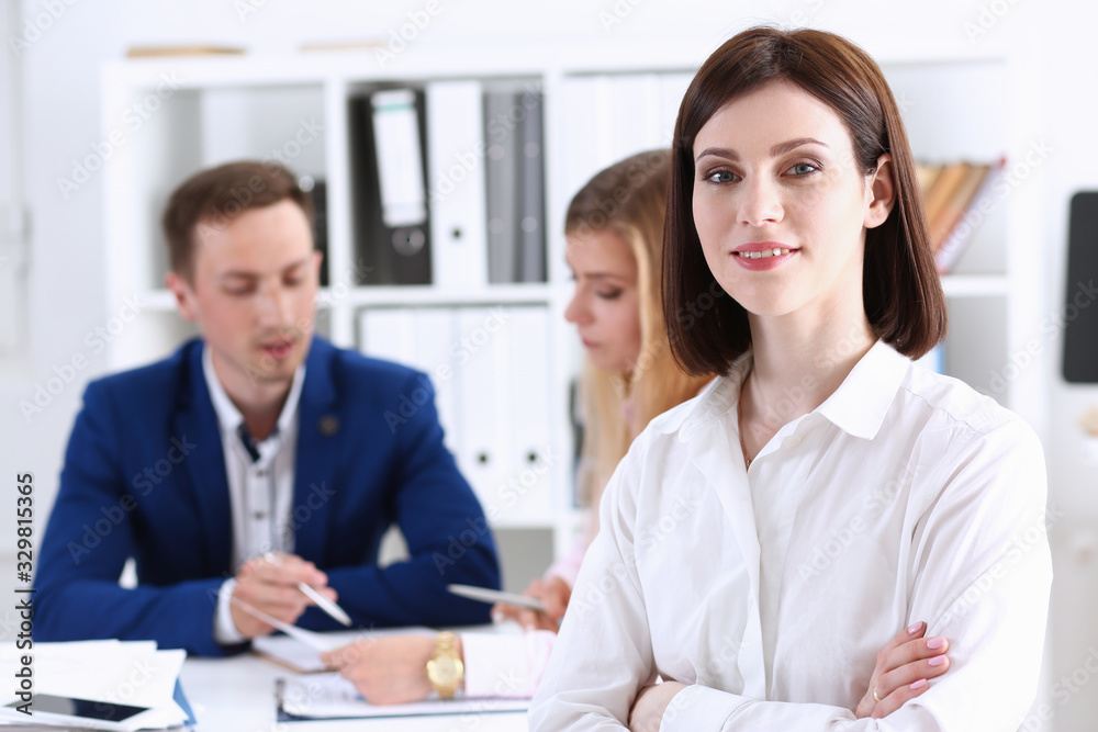 Beautiful smiling cheerful girl at workplace look in camera with colleagues group in background. White collar worker at workspace job offer modern lifestyle client visit profession train concept Beautiful smiling cheerful girl at workplace look in camera with colleagues group in background. White collar worker at workspace job offer modern lifestyle client visit profession train concept