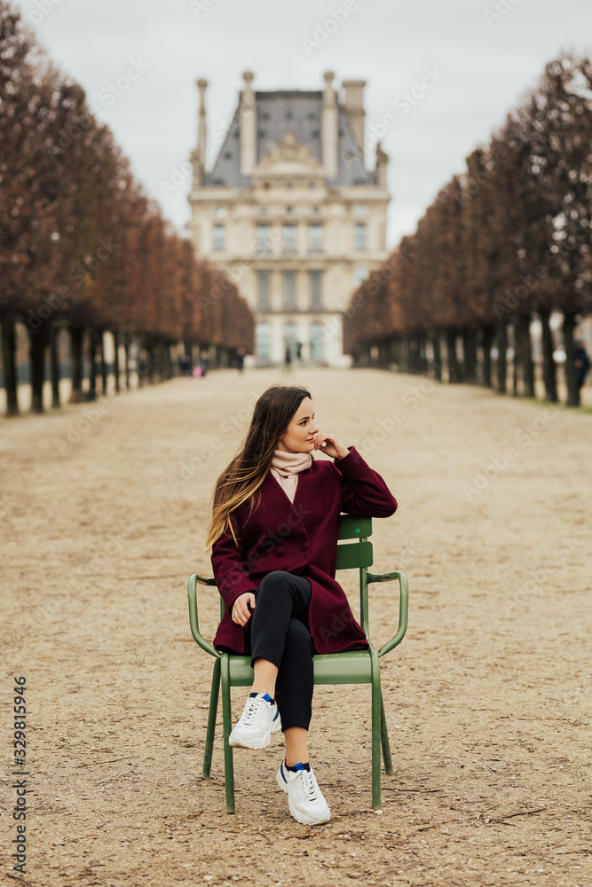 Photo of girl in bordo coat sitting on the chair in front of Louvre Museum in Paris, France. She relaxing and enjoying warm winter day.