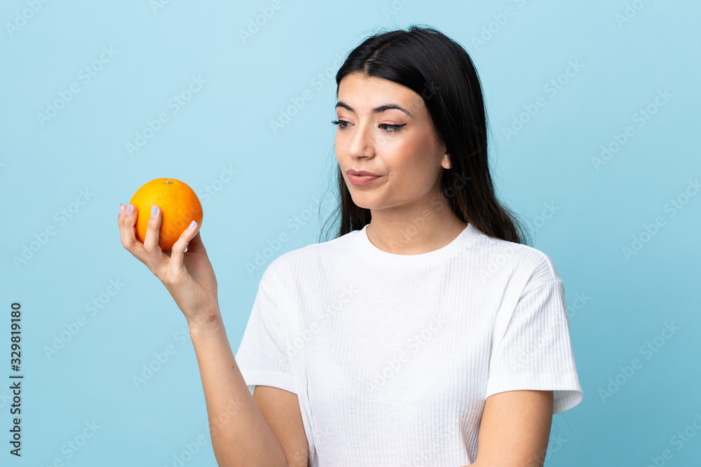 Young brunette girl over isolated blue background holding an orange Young brunette girl over isolated blue background holding an orange