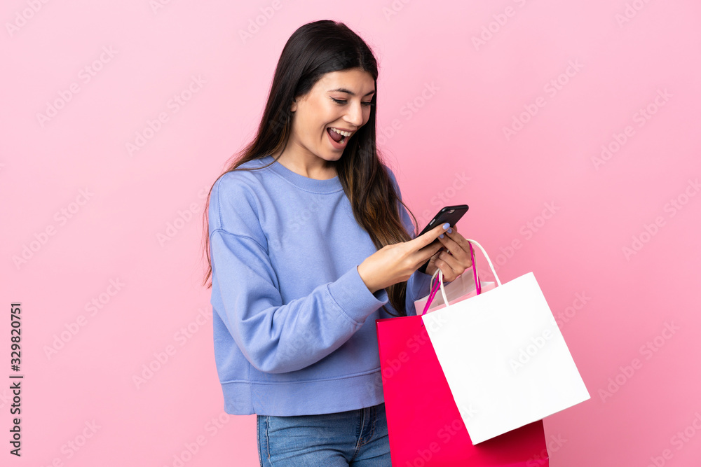 Young brunette girl over isolated pink background holding shopping bags and writing a message with her cell phone to a friend Young brunette girl over isolated pink background holding shopping bags and writing a message with her cell phone to a friend