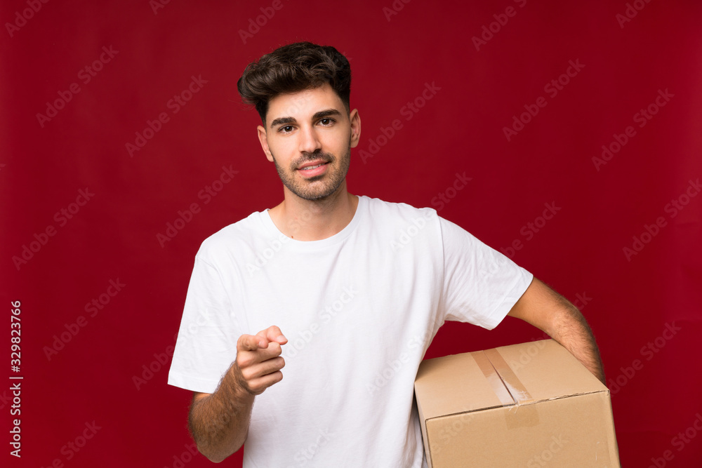 Young man over isolated background holding a box to move it to another site while pointing to the front Young man over isolated background holding a box to move it to another site while pointing to the front