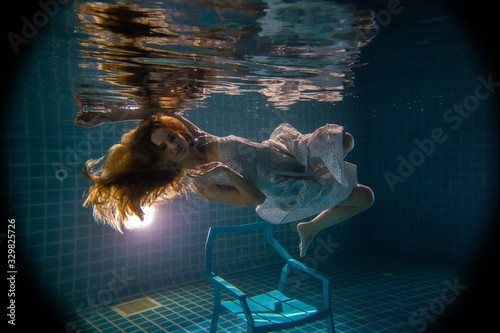 Beautiful woman with long red hair posing underwater on the chair in white dress