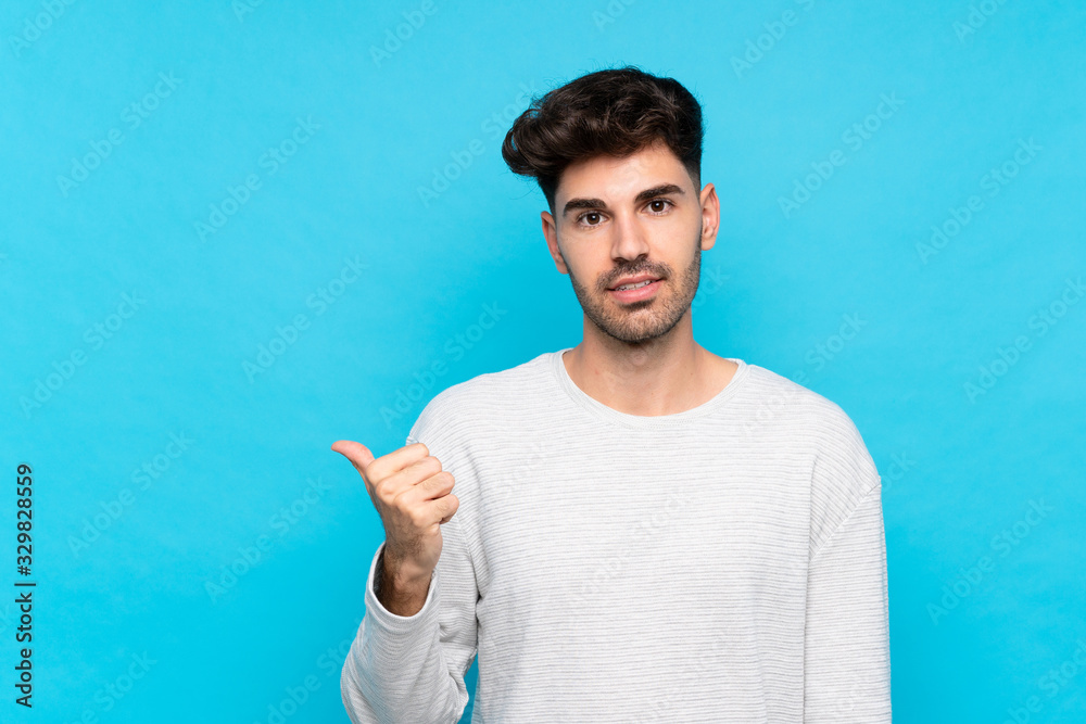 Young man over isolated blue background pointing to the side to present a product Young man over isolated blue background pointing to the side to present a product