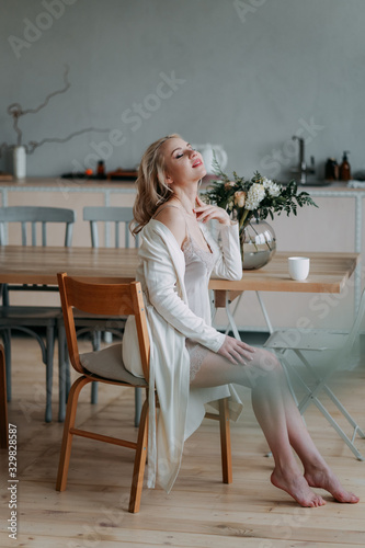 Bride's bouquet. Beautiful happy happy girl blonde bride in a white light sexy robe in the morning in the kitchen is happy, smiling and touching a bouquet of fresh flowers , preparing for the wedding