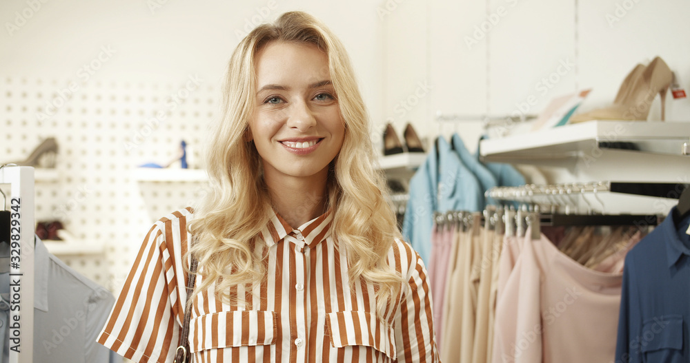 Portrait of young blonde happy woman with curly hair smiling cheerfully to camera in clothing shop. Close up of pretty Caucasian girl with fair hair posing while standing in fancy store. Portrait of young blonde happy woman with curly hair smiling cheerfully to camera in clothing shop. Close up of pretty Caucasian girl with fair hair posing while standing in fancy store.