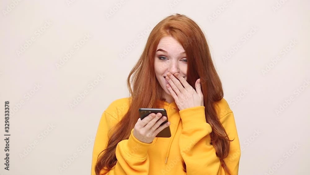 Pretty charming ginger redhead curly young girl in yellow streetwear hoodie isolated on white background in studio. People lifestyle concept. Looking at cellphone using mobile phone chatting have fun