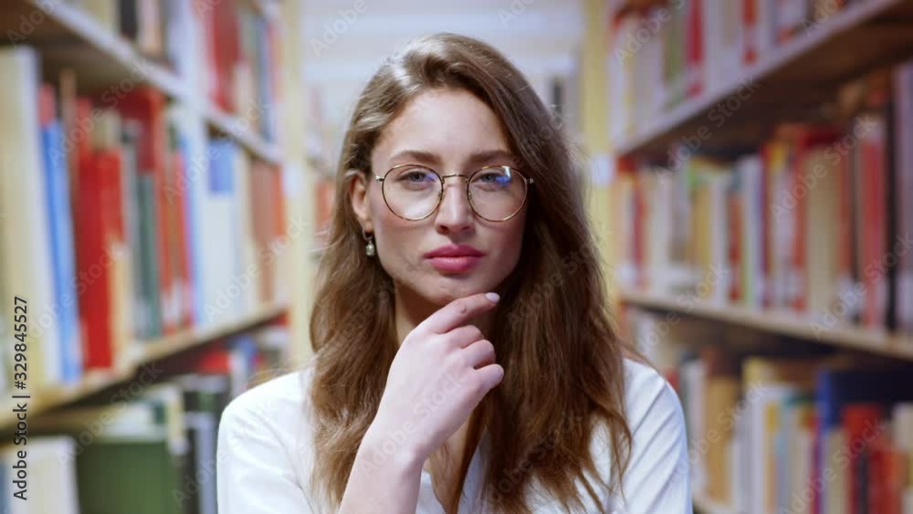Smart serious young pretty female librarian wearing glasses holding her chin thinking looking straight at camera working in the library. Bookshelves college background. Smart serious young pretty female librarian wearing glasses holding her chin thinking looking straight at camera working in the library. Bookshelves college background.