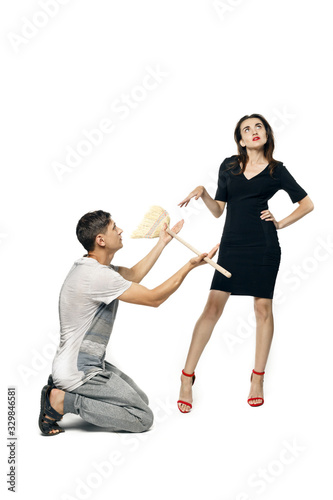 A man in home clothes kneeling holds out a broom to a beautiful woman in evening dress, she dismissively repels him. Isolated on a white background.