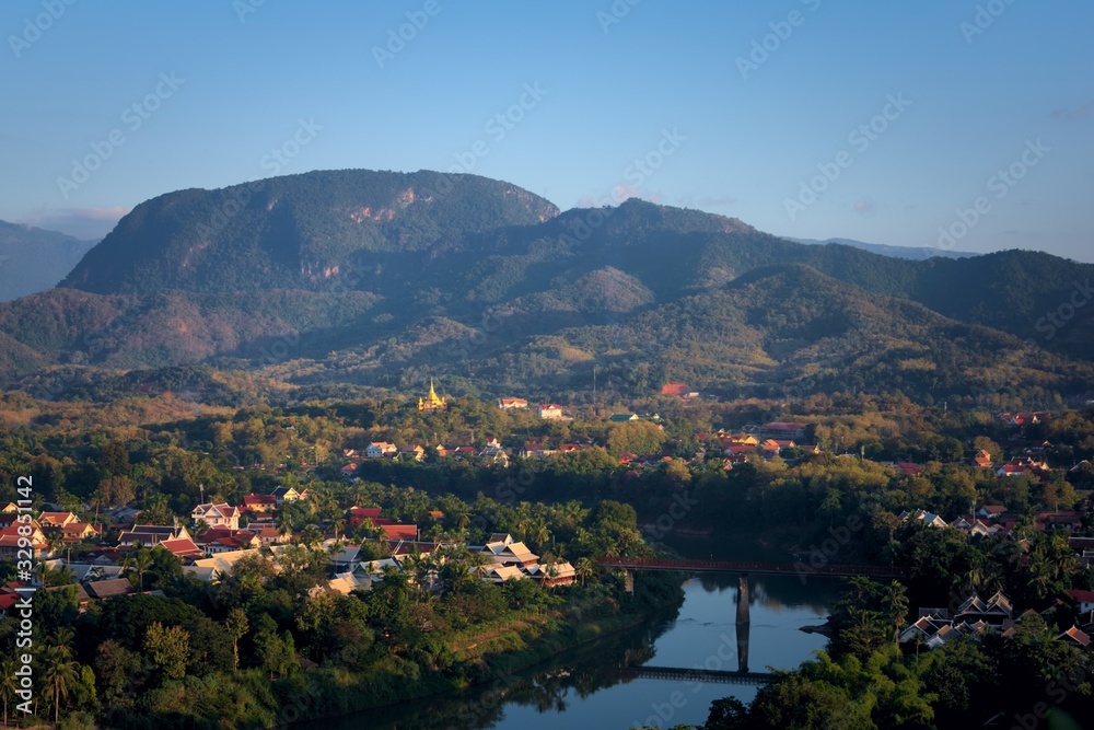 Elevated view of the city of Luang Prabang, Laos, traversed by the Nam Kham river and surrounded by thick rainforest, in the afternoon.