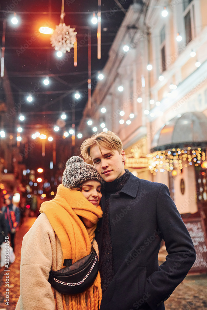 Positive young couple in warm clothes embracing each other on christmas decorated street Positive young couple in warm clothes embracing each other on christmas decorated street