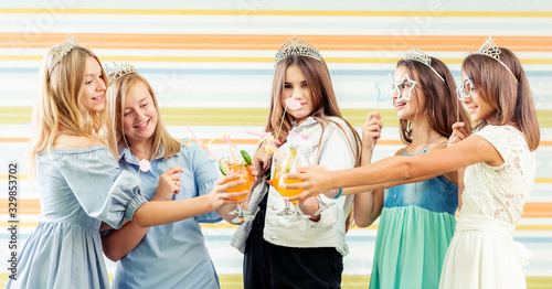 Pretty smiling teenage girls in dresses and crowns knocking beverages together at birthday party