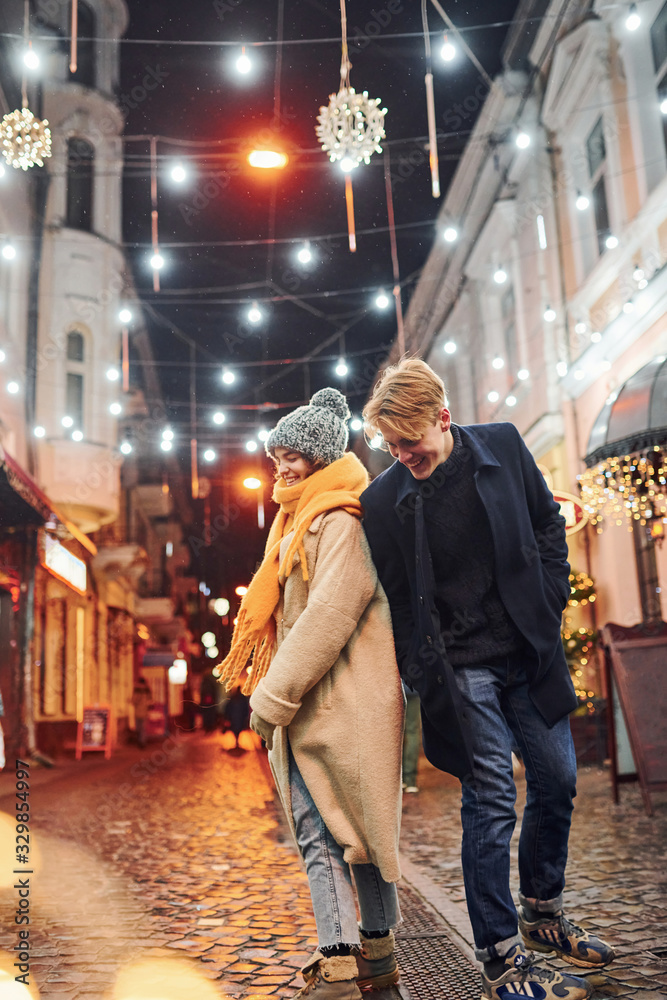 Cute couple in warm clothes is on christmas decorated street smiling and have fun