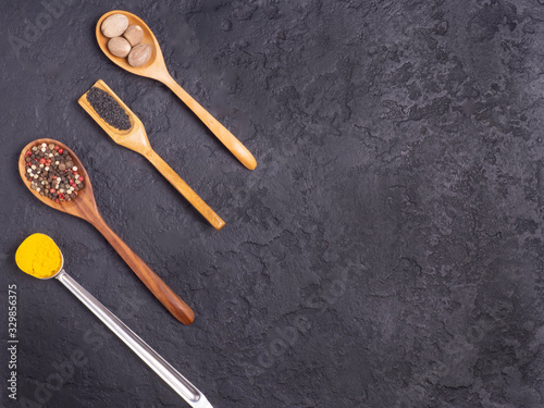 Different spices in wooden and metal spoons on a black concrete background.