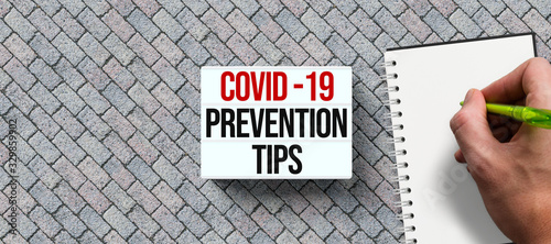 Plakat lightbox with text COVID-19 PREVENTION TIPS and hand with pen over a notepad on stone background