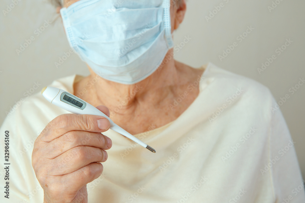 An elderly woman with a medical mask on her face is holding a thermometer with a high temperature An elderly woman with a medical mask on her face is holding a thermometer with a high temperature