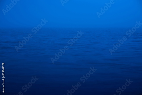 Water ripple nature background. Natural texture of sea of blue classic color. Deep blue calm water in dusk close-up. Meditative image of dense fog above lake. Soft light glitters on water surface. © Daniil