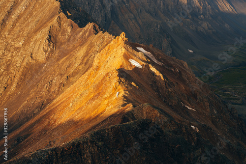 Atmospheric alpine landscape with shiny rockies with snow in golden hour. Scenic view to big orange rocks in sunrise. Wonderful highland scenery with gold pointy peak with firn. Flying over mountains.