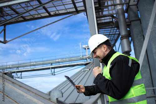 Factory worker standing on metal platform above industrial storage tanks and checking production results on his tablet computer.