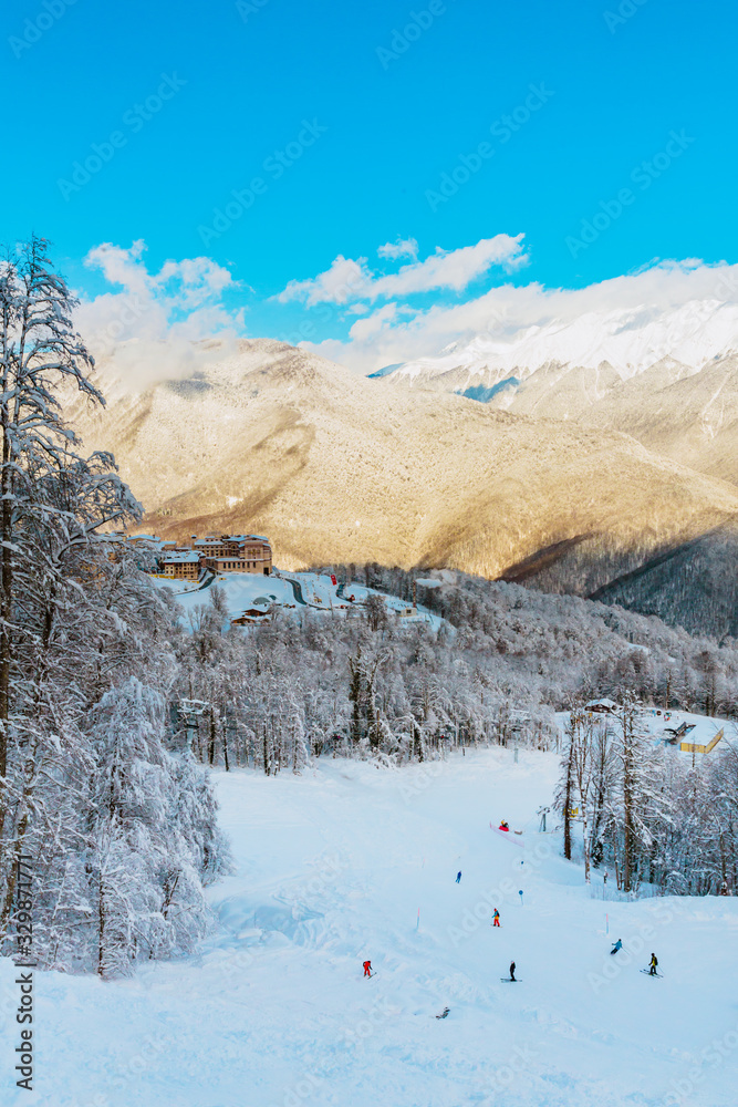 Ski resort panorama with skiing people, ski and snowboard riders, snowy forest around, buildings backwards, view on high snowy rocky mountains in sunny day and blue sky. Vertical image