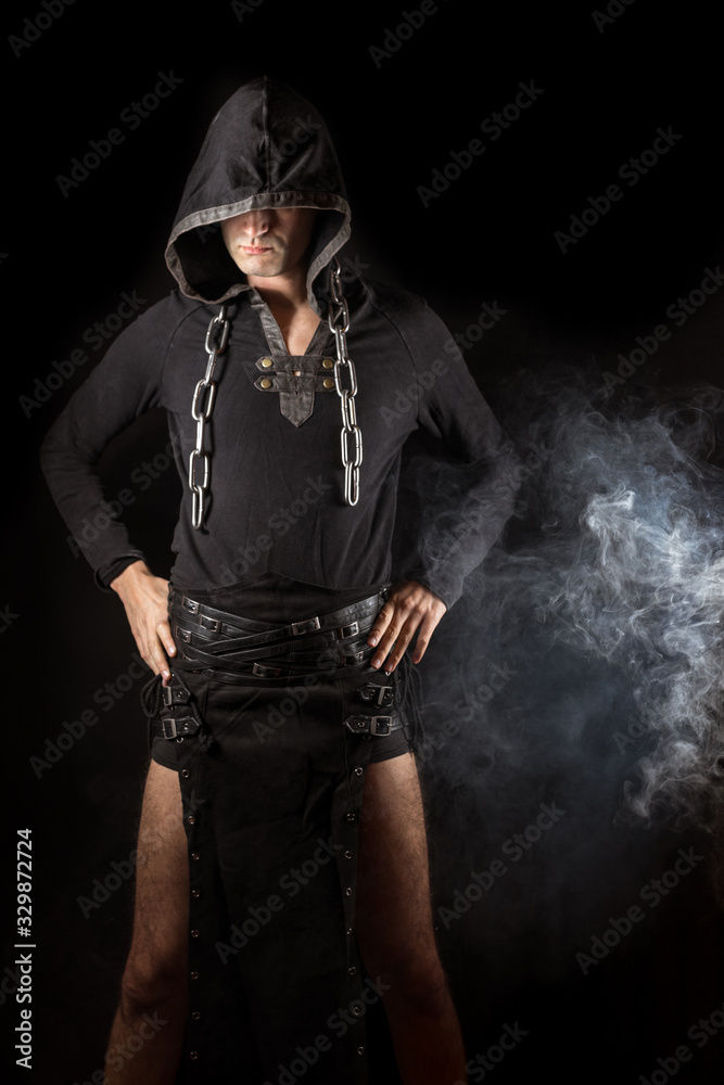 Young man posing in hooded gothic clothes with chain around neck next to smoke Young man posing in hooded gothic clothes with chain around neck next to smoke