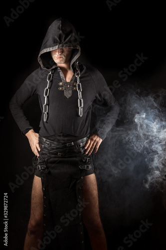 Young man posing in hooded gothic clothes with chain around neck next to smoke