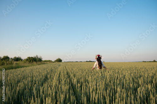 Pretty young woman wear traditional Ukrainian clothes and flower wreath walk in wheat field, beautiful ethnic girl in handmade decorated floral crown admire nature, blue sky background © Kokhan O