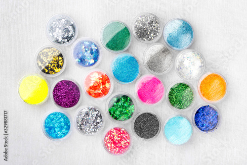 Creative flat lay overhead composition with sequins, glitter, mica and shimmer for nail design in plastic jars on a light wooden background. Top view.