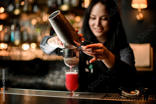 smiling woman bartender pours cocktail from shaker into glass using sieve.