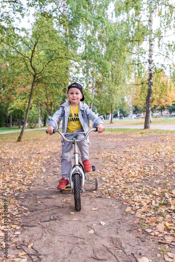 Little boy 3-5 years old, summer park, background trees. Trains learns keep balance bicycle, rides pedals. In summer in city, emotions interest comfort happiness. Casual wear, denim baseball cap. Little boy 3-5 years old, summer park, background trees. Trains learns keep balance bicycle, rides pedals. In summer in city, emotions interest comfort happiness. Casual wear, denim baseball cap.