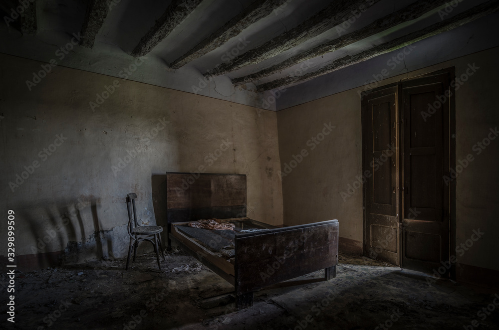 Dormitorio de casa abandonada con cama y silla en la penumbra iluminada por una ventana.
