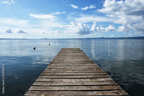 Wooden jetty for boats against a blue sky with some clouds. Floating buoys. Hills on background. Bolsena lake, Italy.