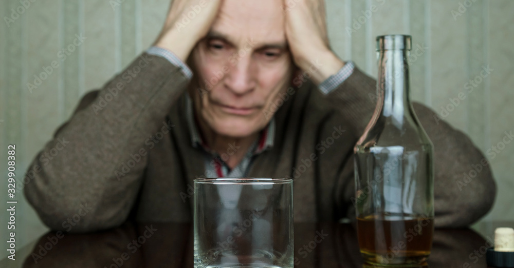 depressed senior man sits at brown table looking at bottle with whiskey and empty glass