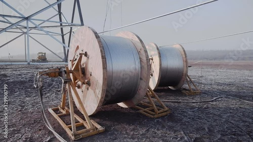 large wooden bobbins with uncoiling high-voltage wires near power transmission tower with special equipment on ground