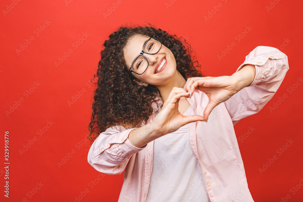 Naklejka premium Portrait of a smiling young curly woman showing heart gesture with two hands and looking at camera isolated over red background.