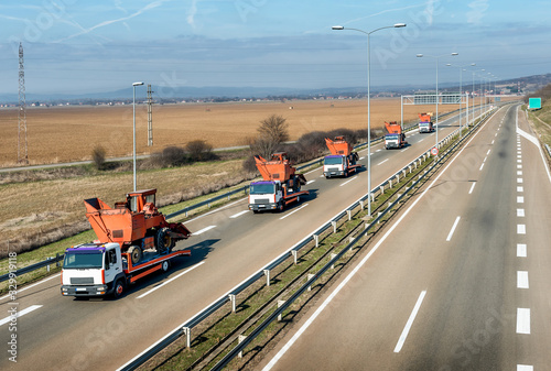 Tow trucks or Flatbed trucks in a convoy towing agricultural or construction machines under a beautiful sky on a highway