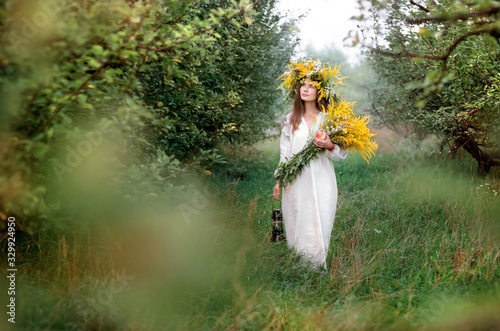 Young beautiful woman in a wreath of wildflowers and a long white national shirt walks in the apple orchard. Ukrainian culture, ethno style