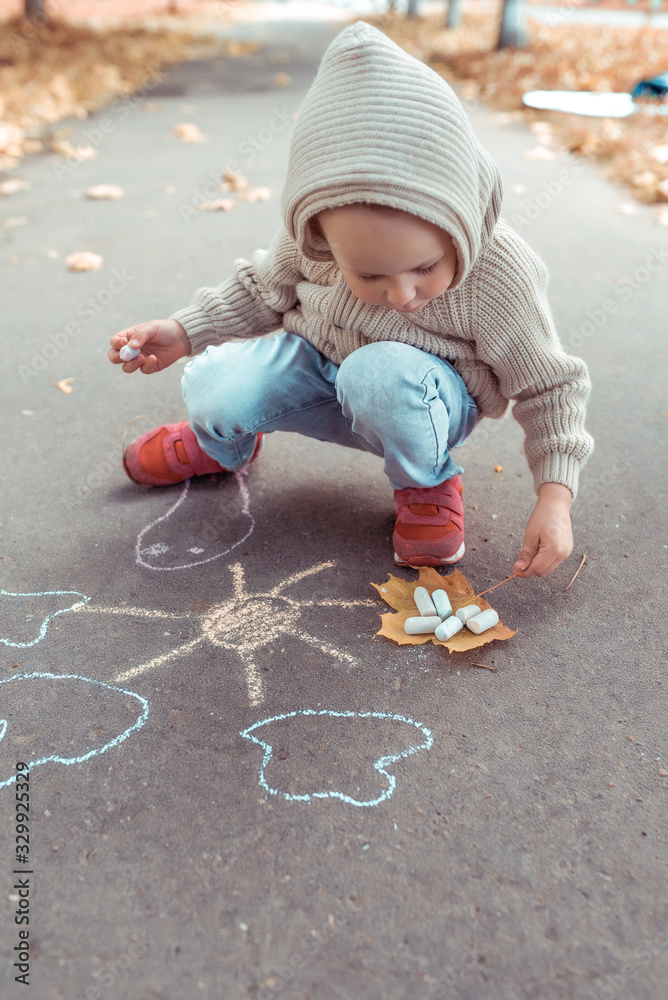 Fototapeta premium little boy in beige jumper with hood, autumn day in summer in city, draws sun on asphalt with colorful chalks. Creative concept idea and creativity.